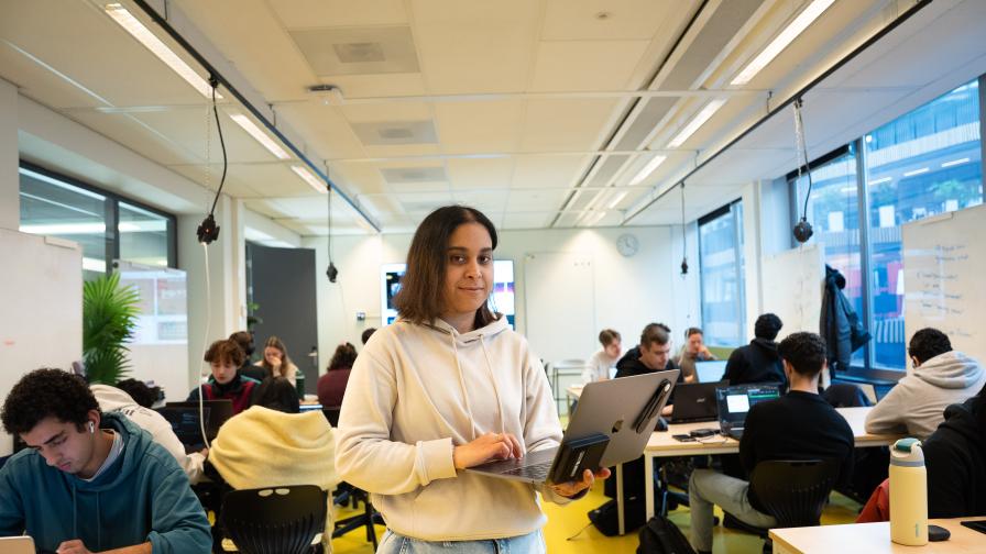 Frontend Design en Development student Chama staat met laptop in de handen met klasgenoten op de achtergrond aan tafelhter laptop