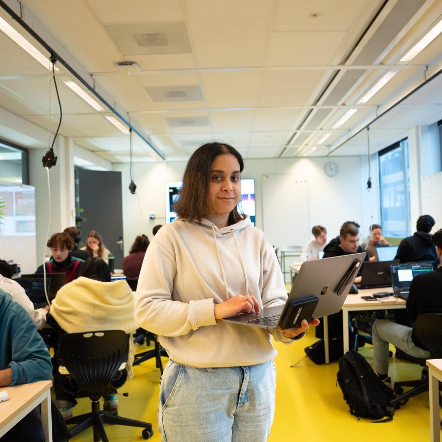 Frontend Design en Development student Chama staat met laptop in de handen met klasgenoten op de achtergrond aan tafelhter laptop
